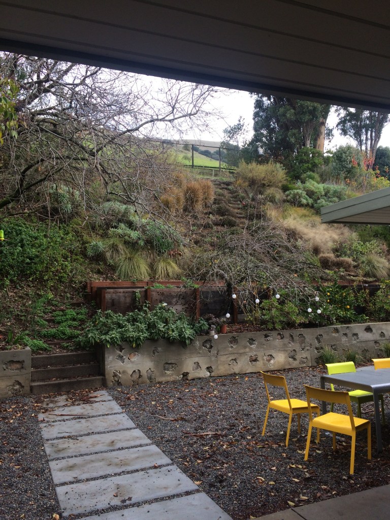 A view of a landscaped outdoor area featuring stone steps, greenery, and a gravel surface. Bright yellow chairs surround a table, while a wooden structure is visible in the background.