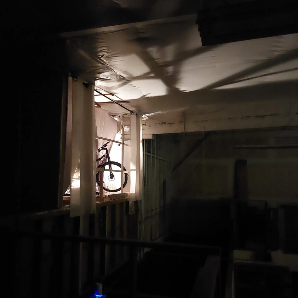 A dimly lit interior space with a bicycle positioned near a window, casting shadows on the walls and ceiling.