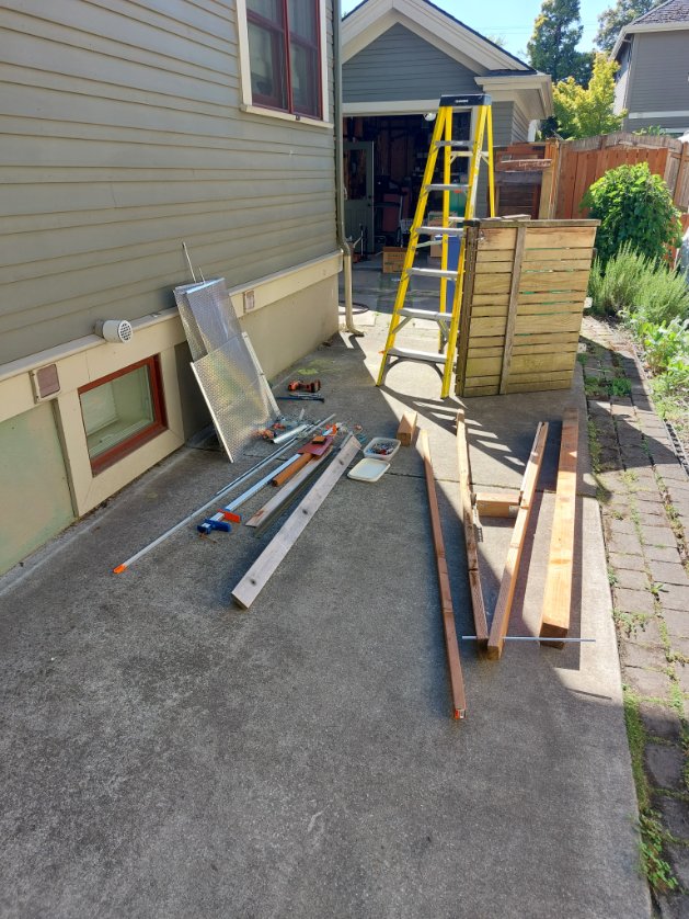 A backyard area with a yellow ladder, wooden planks, and various tools laid out on concrete. A wooden structure and a window of a house are visible in the background.